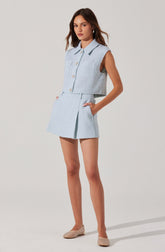A young woman is wearing a light blue sleeveless button-up vest with a collar and large buttons down the front. She pairs it with matching high-waisted shorts that have pleats and pockets. She completes the outfit with beige textured flat shoes.